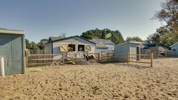 A stable with a horse, surrounded by wooden fences and sheds in a sandy area.