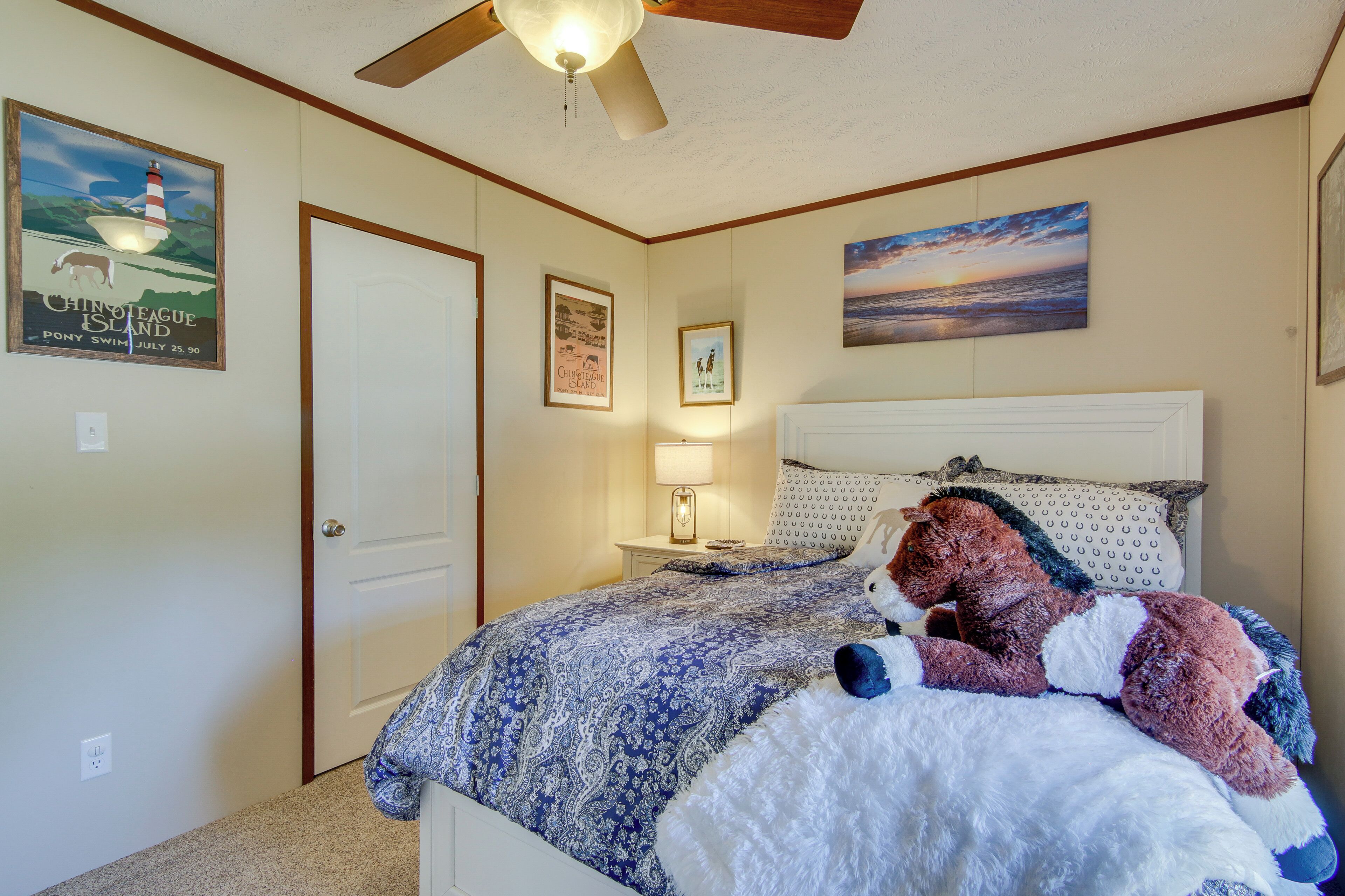 A bedroom with a bed, a ceiling fan, a nightstand with a lamp, and wall decorations including a poster and a painting.