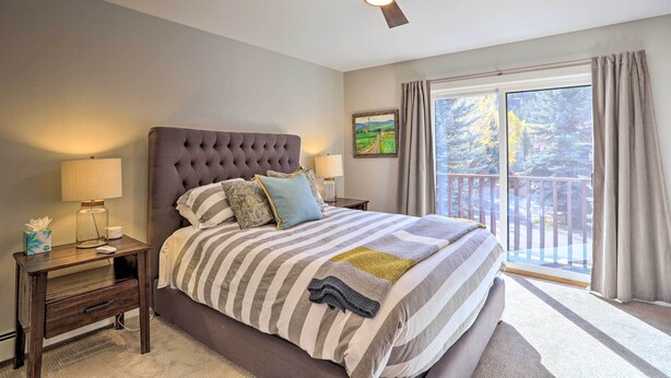 Een slaapkamer met een groot bed, een plafondventilator, een nachtkastje met een lamp en een raam met uitzicht op bomen.
