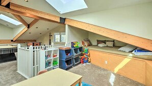 Een ruime kamer met een schuine kap, houten balken, een stapelbed, een speelruimte met speelgoed en een tafel.