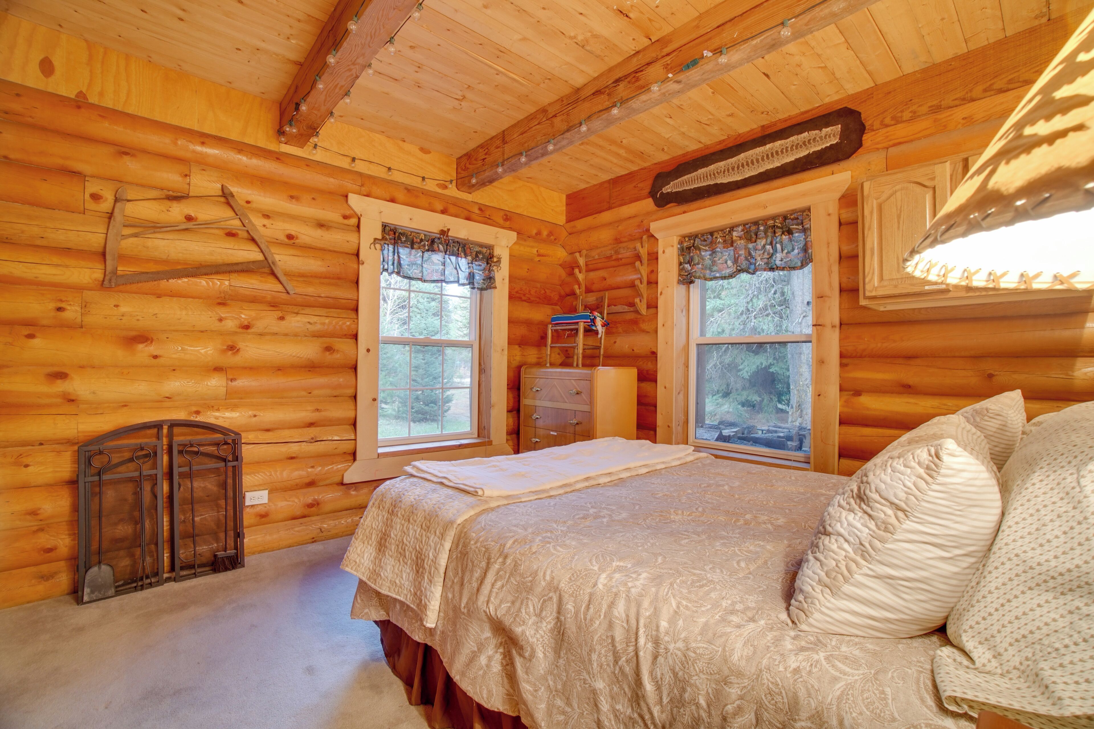 Quarto de cabana de tronco com uma cama, duas janelas com cortinas, uma escada e uma cômoda pequena.