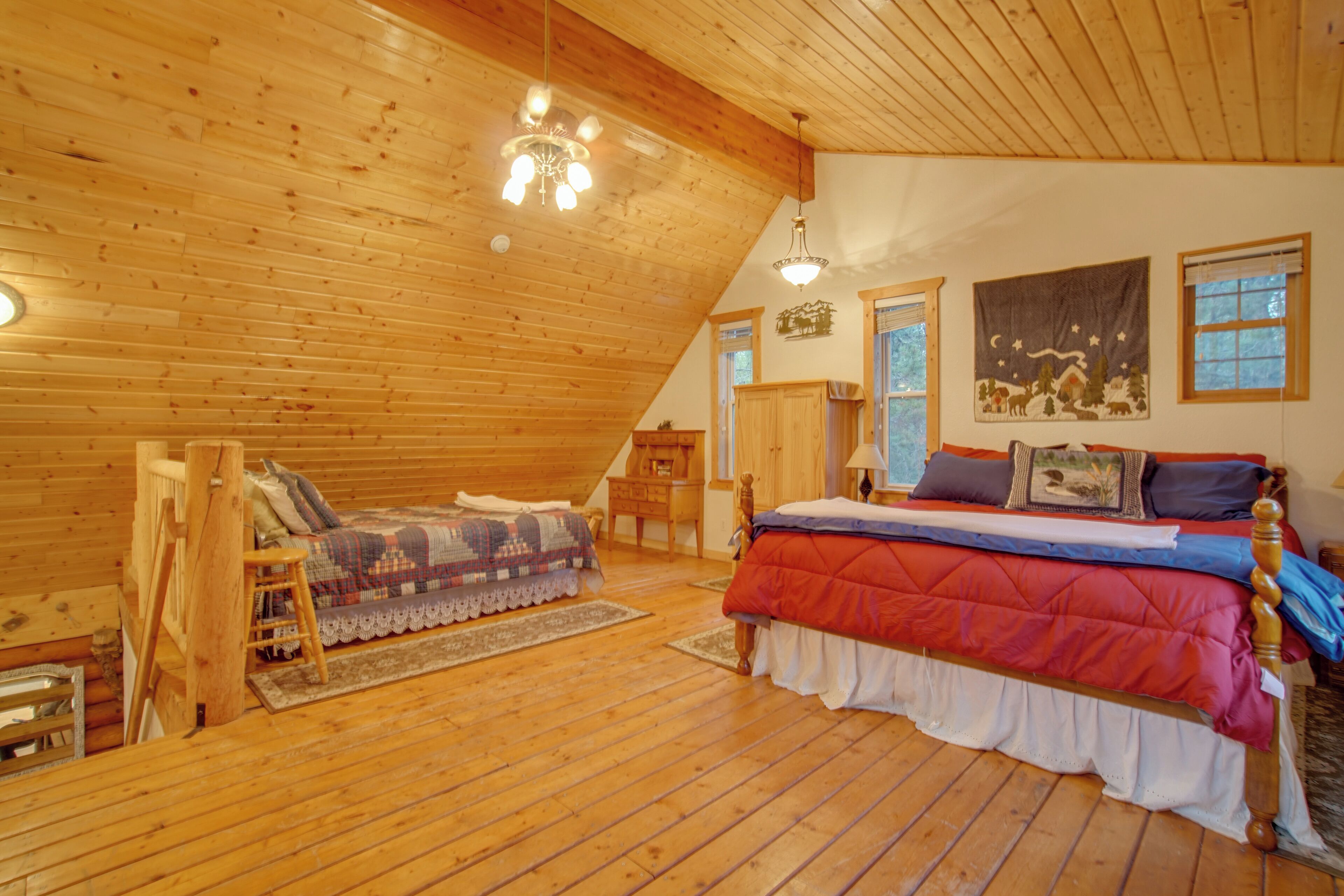 A cozy attic bedroom with a wooden ceiling, a large bed with a red and blue quilt, a wooden ladder, and a small desk.