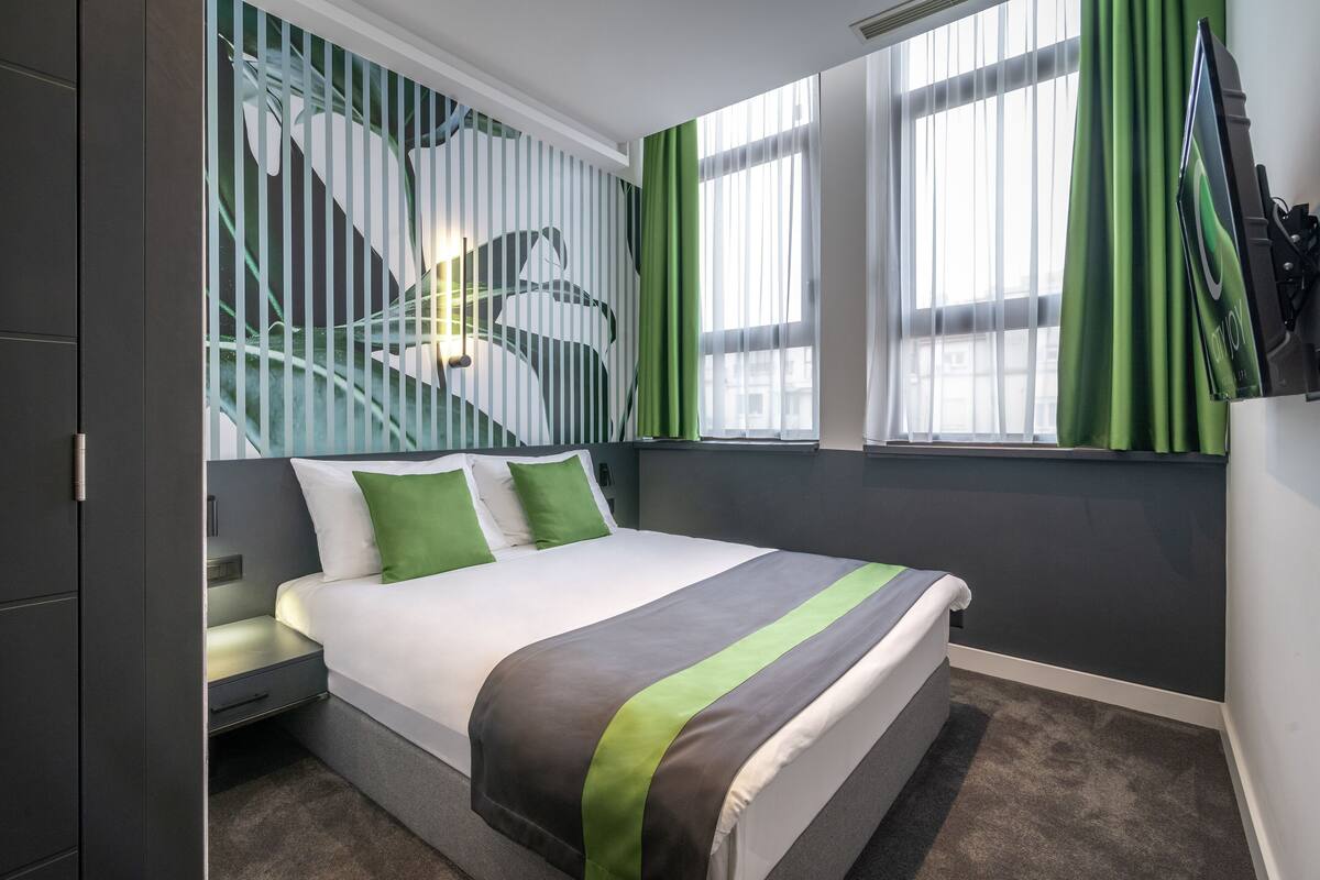 Quarto de hotel moderno com uma cama grande, cortinas verdes e um mural na parede.