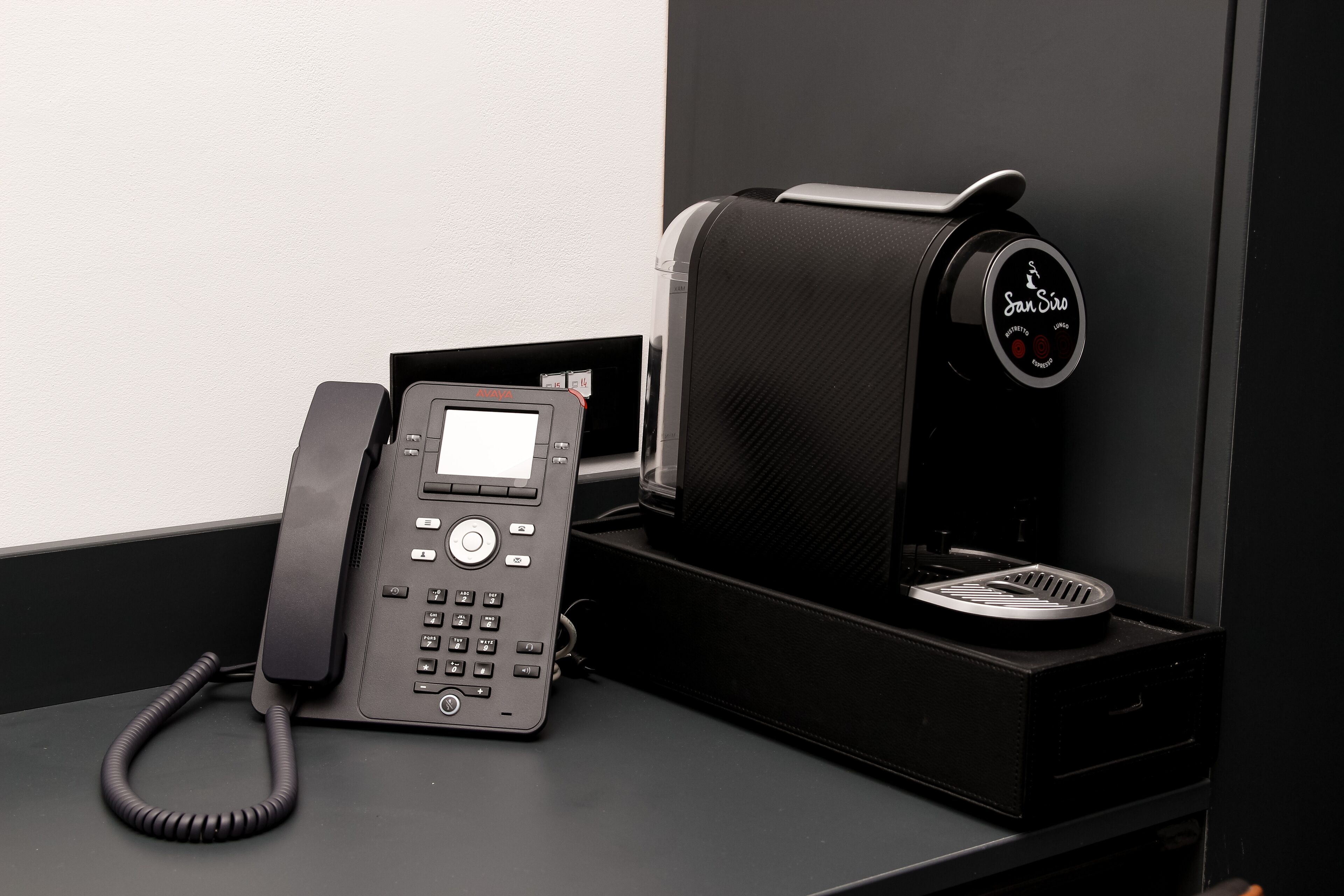 A black telephone on a desk with a coffee machine beside it.