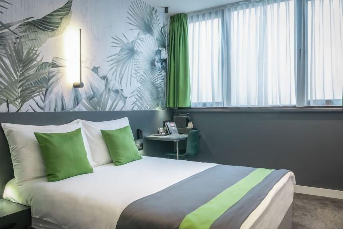 Habitación de hotel con cama, almohadas verdes, escritorio con teléfono y ventana con cortinas de gasa.