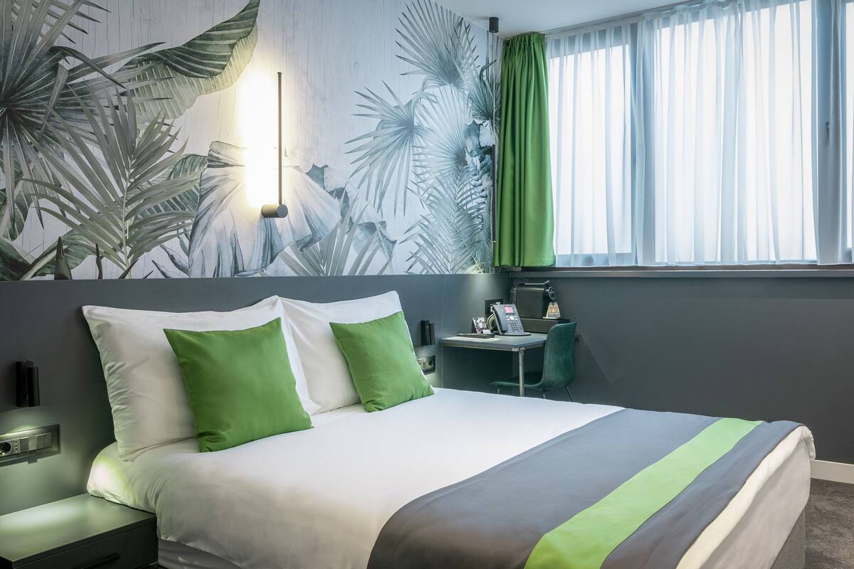 Quarto de hotel com cama, escrivaninha com telefone e janela com cortinas verdes.