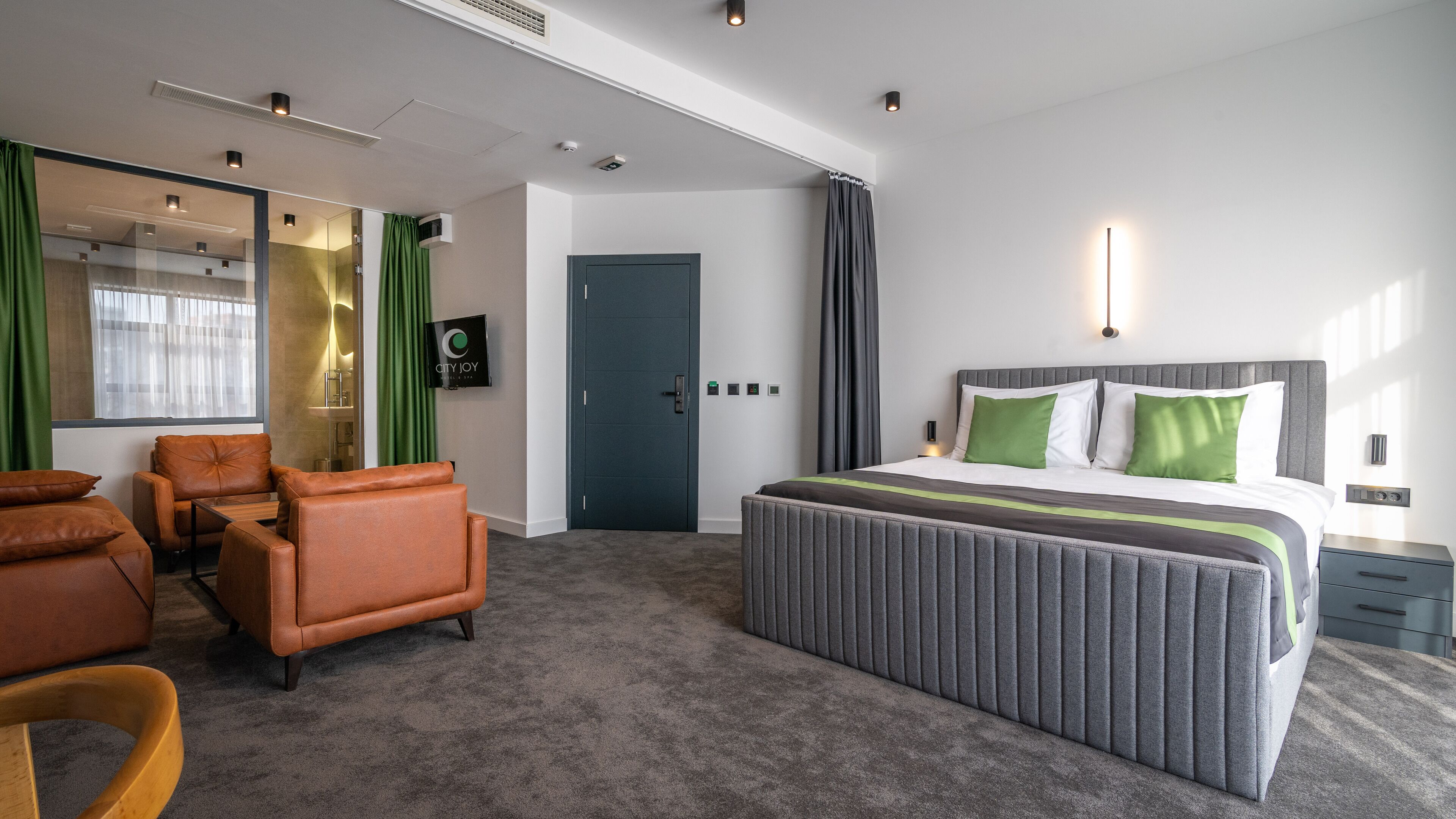 Een moderne hotelkamer met een groot bed, een zithoek met lederen fauteuils, een flatscreen televisie en een spiegel.