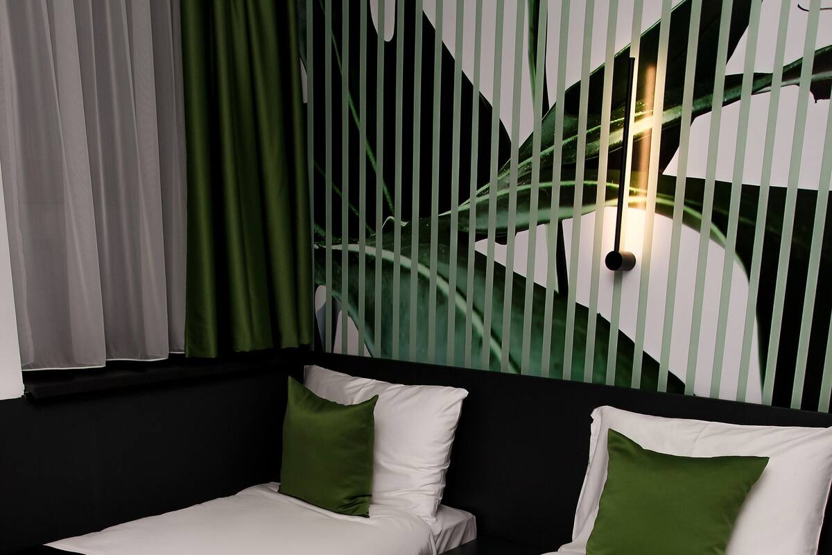 Quarto com duas camas, cada uma com roupa de cama branca e verde, cabeceira preta e cortina verde.