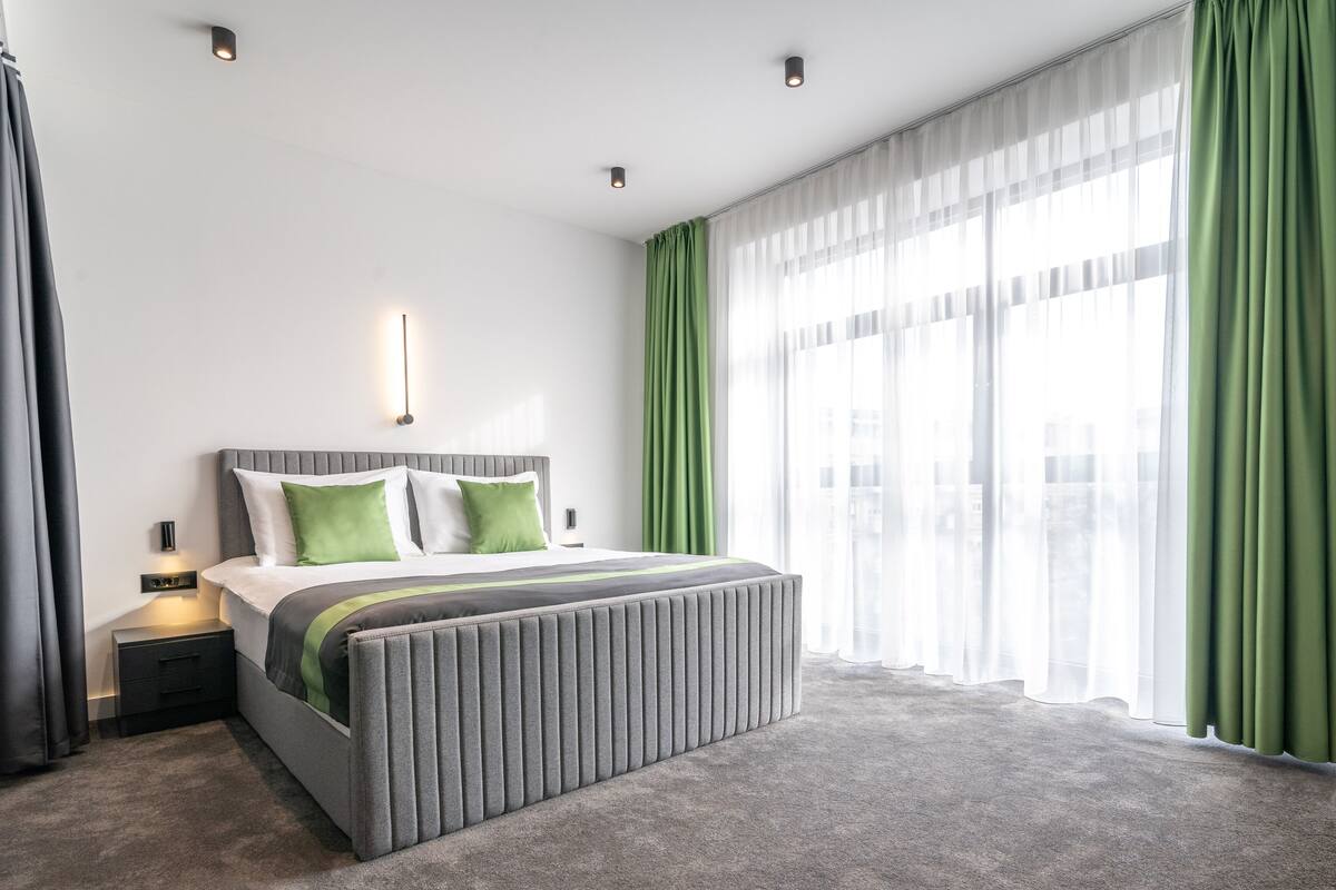 Quarto de hotel moderno com uma cama grande, cortinas verdes e vista para a cidade.