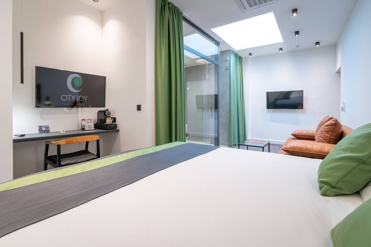 Quarto de hotel moderno com cama, escrivaninha com computador, televisão e chuveiro de vidro.