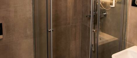 Cabina de ducha con paredes de vidrio, ducha de mano, puerta de vidrio y un lavamanos blanco a la derecha.
