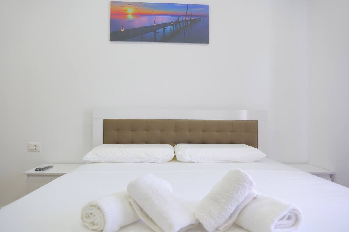 A hotel room with a bed, white linens, a headboard, a painting of a sunset over a pier, and a bedside table with a remote control.