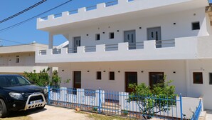 Un bâtiment blanc à deux étages, avec une clôture bleue, une voiture garée et de petits arbres dans le jardin.