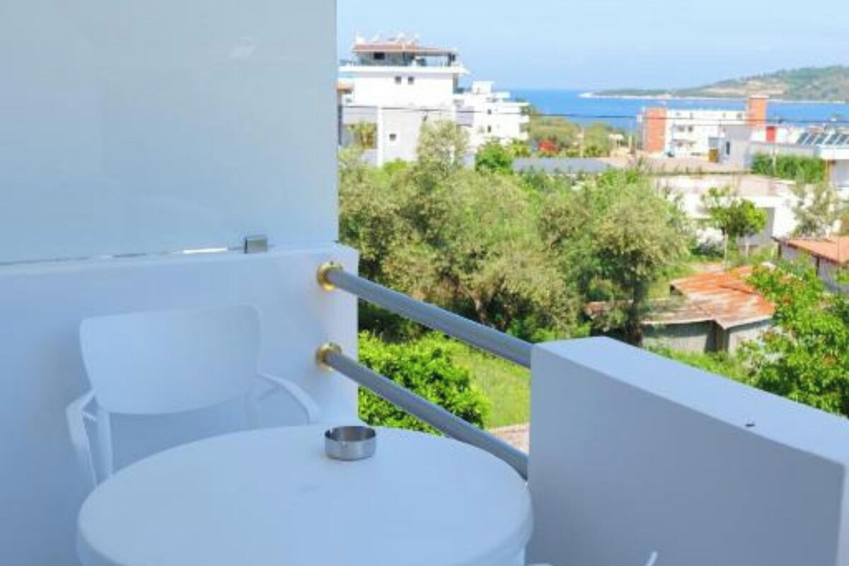 A balcony with a white table and chairs, overlooking a coastal area with buildings and greenery.
