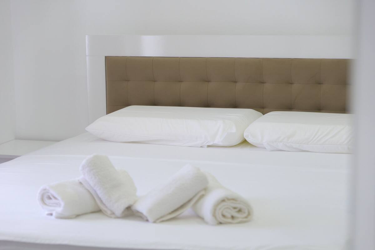 A neatly made bed with white linens and a beige headboard.