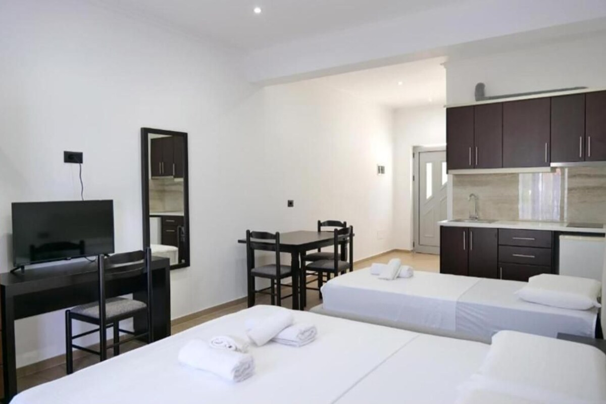 A hotel room with two beds, a desk with a TV, a dining table, and a kitchenette.