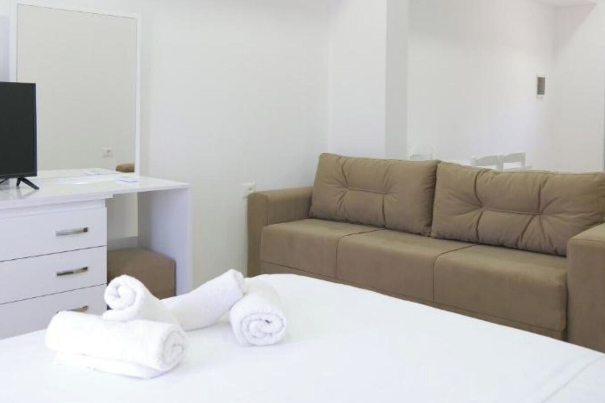 A hotel room with a bed, a white bedside table with a flat-screen TV, a beige sofa, and rolled white towels.