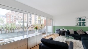 A modern living room with a large window, a black sofa, a glass coffee table, and a white rug. - Cosy 1bedroom Flat - 2 Min Walk to Kensington Gardens! (London)