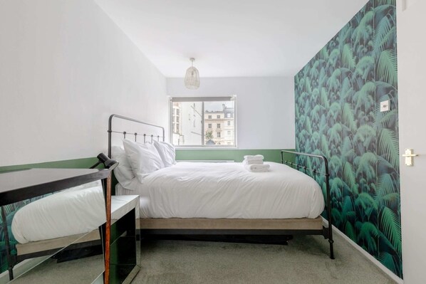 A bedroom with a bunk bed, a single bed, a window with a view of buildings, and a green leafy wallpaper.