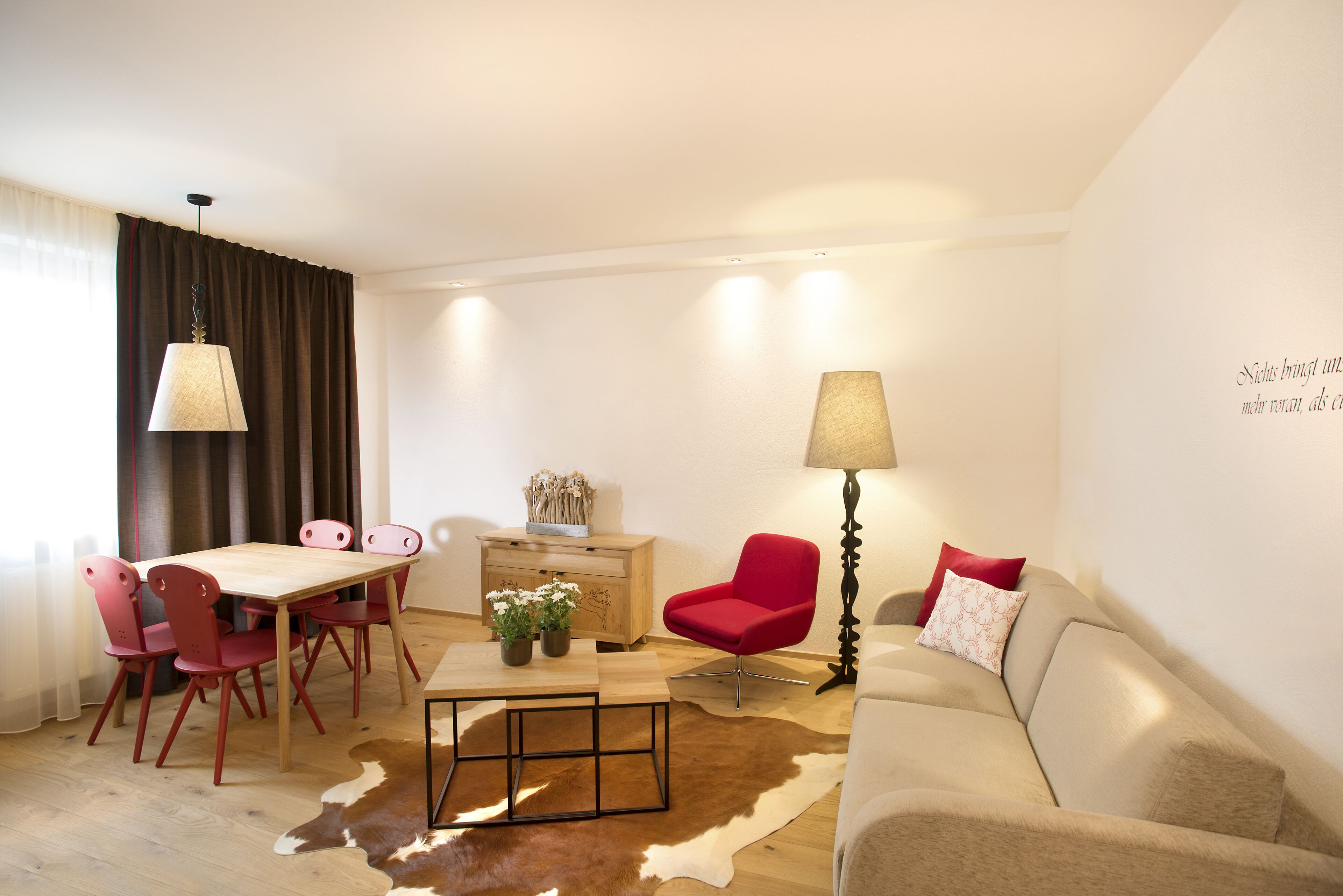 En moderne stue med et træspisebord, røde stole, en beige sofa og en rug af oksehud.