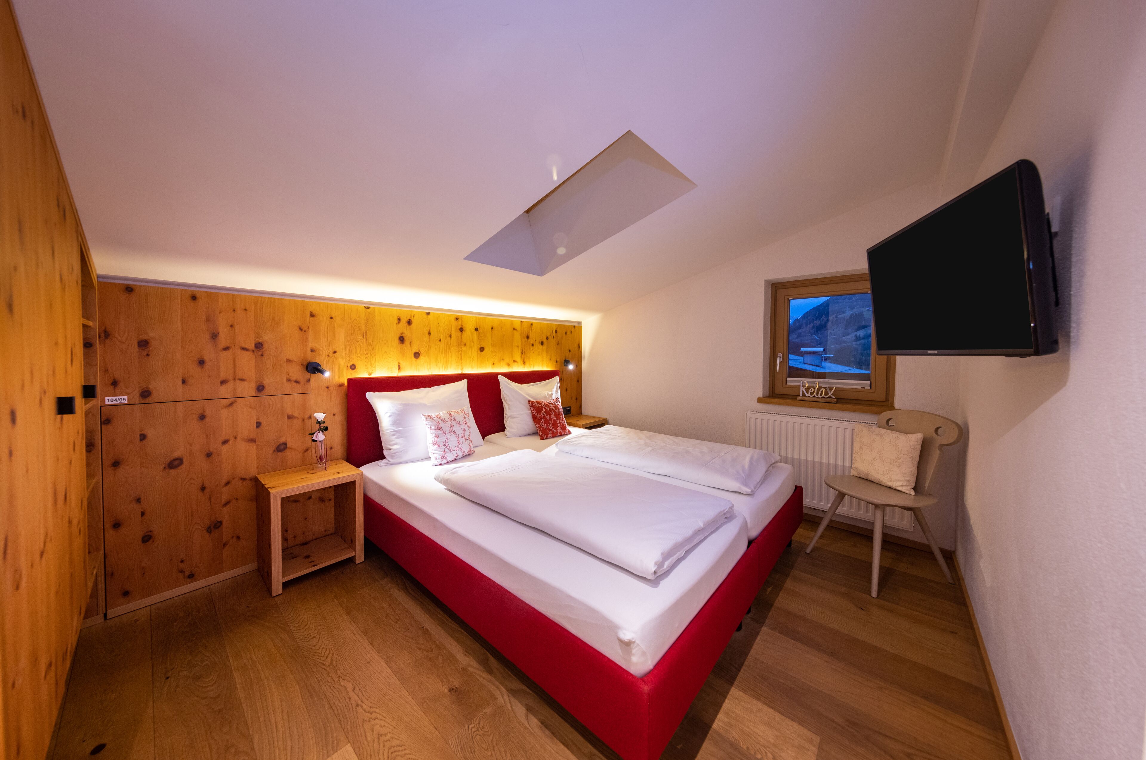 Ein Hotelzimmer mit einem hölzernen Kopfteil, einem Bett mit weißer Bettwäsche, einem Nachttisch, einem Sessel, einem an der Wand befestigten Fernseher und einem Oberlicht.