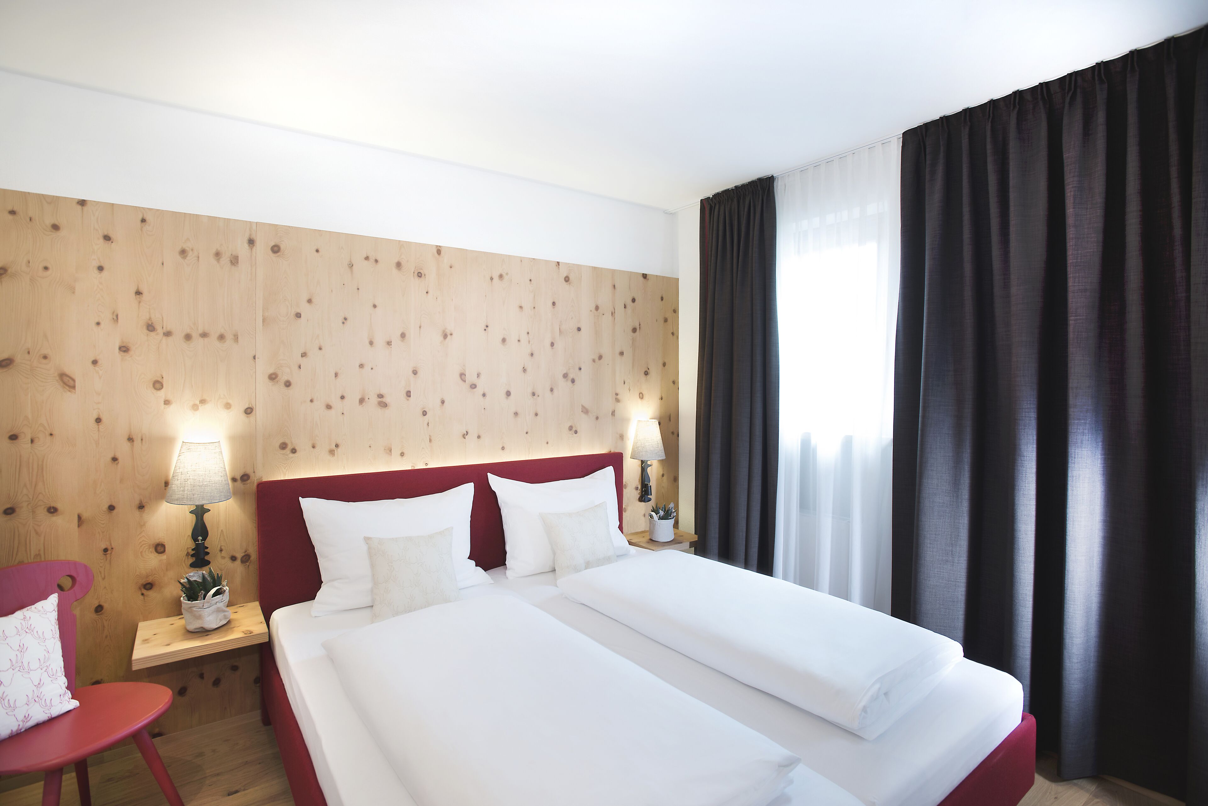Ein Hotelzimmer mit zwei Betten, Holzwandverkleidung, einem roten Kopfteil, einem Nachttisch mit Lampe, einem roten Sessel und einem Fenster mit dunklen Vorhängen.