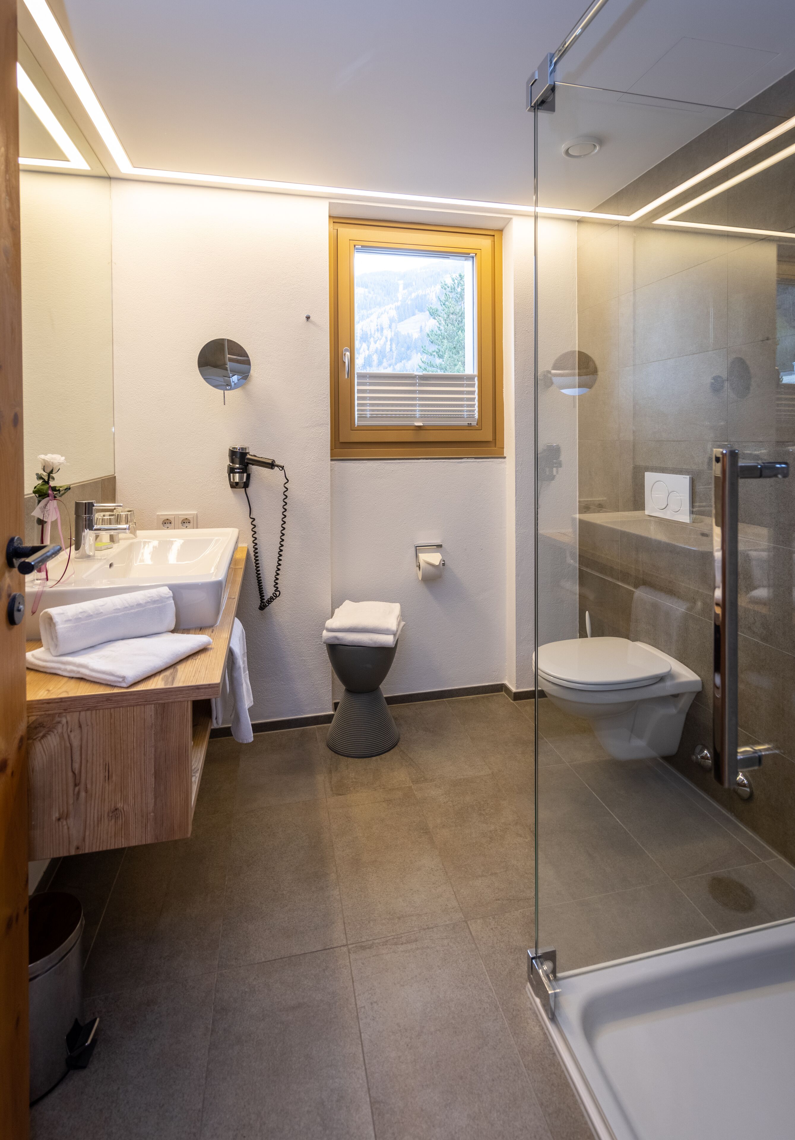 Un baño moderno con cabina de ducha de vidrio, tocador de madera, inodoro y ventana que deja entrar luz natural.