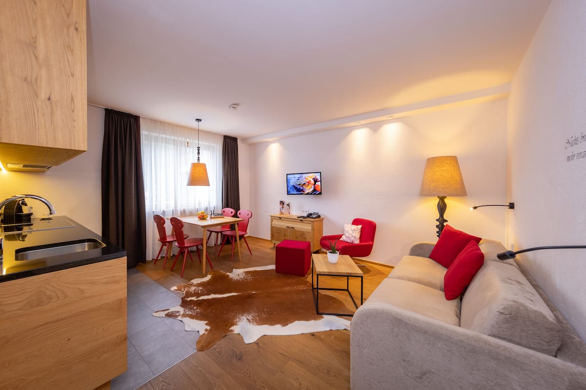 A modern living room with a sofa, a dining area with chairs, a kitchen with a sink, and a TV mounted on the wall.