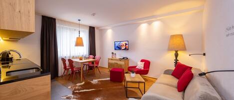 A modern living room with a sofa, a dining area with chairs, a kitchen with a sink, and a TV mounted on the wall.