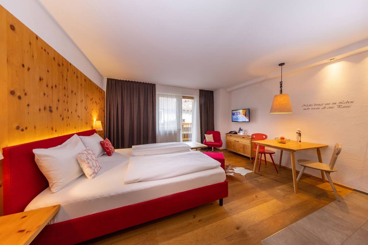 A hotel room with a red sofa bed, a wooden headboard, a desk with a chair, a television, and a wall-mounted lamp.
