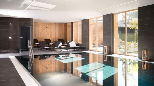 A modern indoor swimming pool with a glass enclosure, wooden panel walls, and a seating area with lounge chairs. - Hapimag Ferienwohnungen Zell am See (Maishofen)