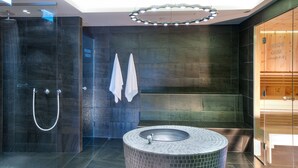 A modern bathroom with a glass-enclosed shower, a round sink with mosaic tiles, and a sauna visible through a wooden door. - Hapimag Ferienwohnungen Zell am See (Maishofen)