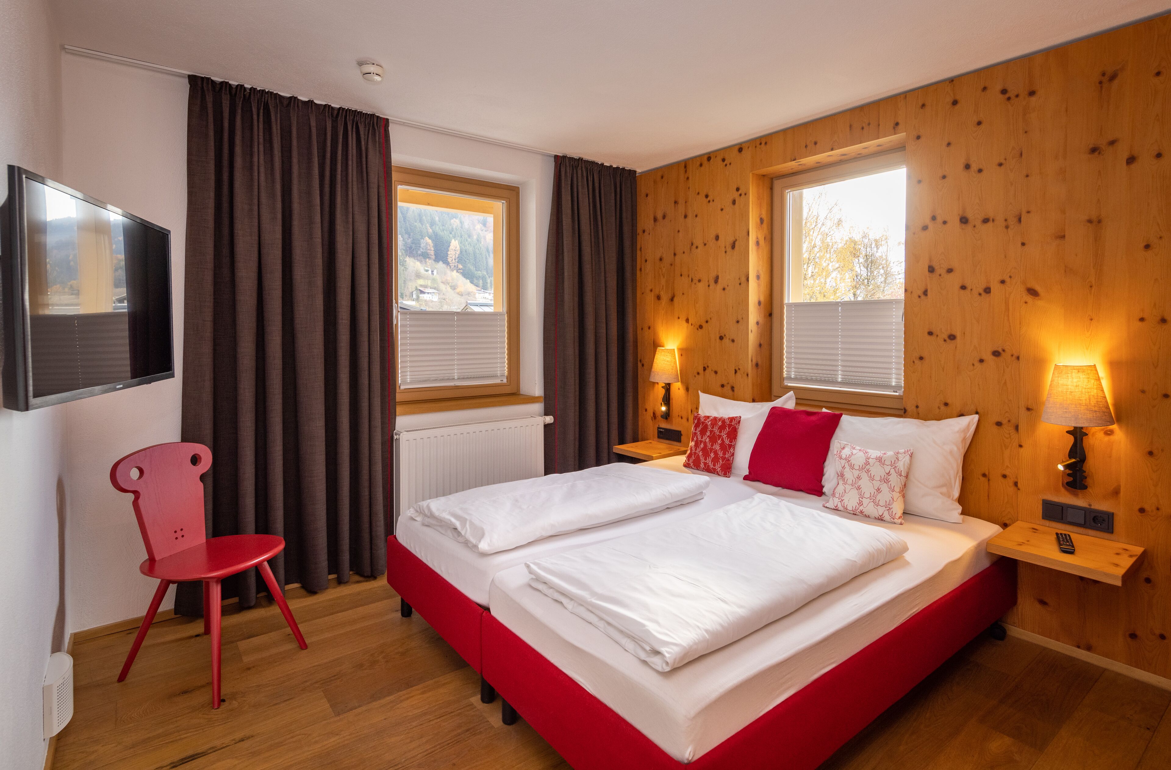 A double bed with white bedding, a red chair, two bedside tables with lamps, and a TV on the wall.