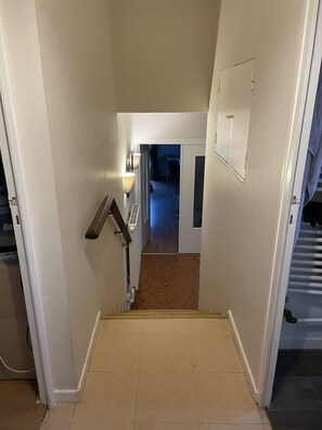 Un escalier étroit doté d’une main courante, un luminaire fixé au mur et une porte en haut, donnant sur une pièce au plancher sombre.