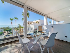 Un espace repas moderne avec une table en verre et des chaises blanches, donnant sur un paysage urbain avec des palmiers et des immeubles.
