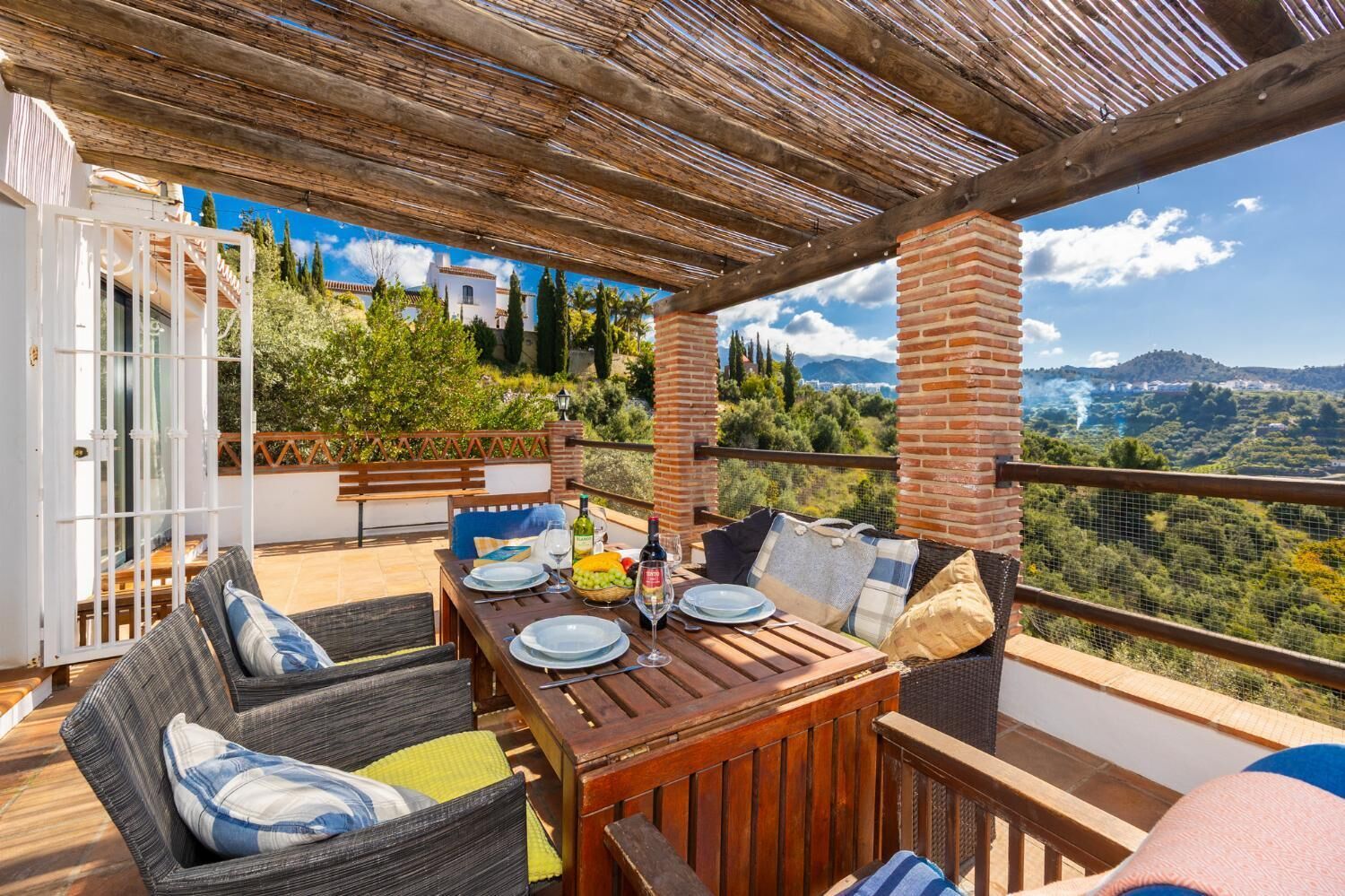 A terrace with a table set for dining, wicker furniture, and a view of a mountainous landscape.