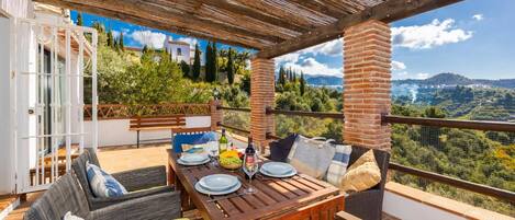 A terrace with a table set for dining, wicker furniture, and a view of a mountainous landscape.