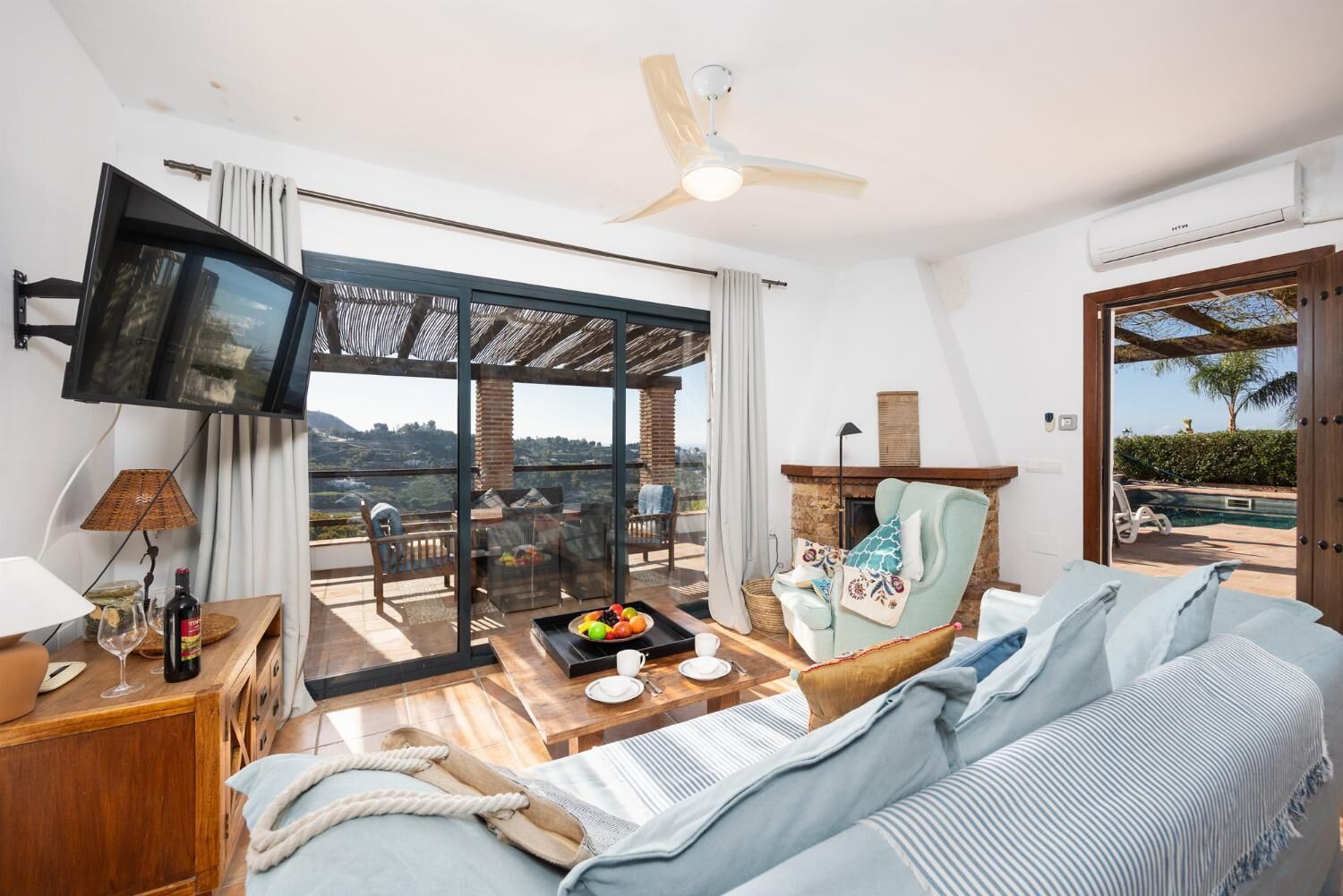 Een moderne woonkamer met een bank, een televisie, een eettafel met een schaal fruit, en een balkon met uitzicht.