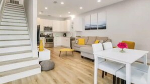 Un apartamento moderno con una escalera, una sala de estar con sofá y mesa de centro, un comedor con mesa y sillas, y una cocina al fondo.