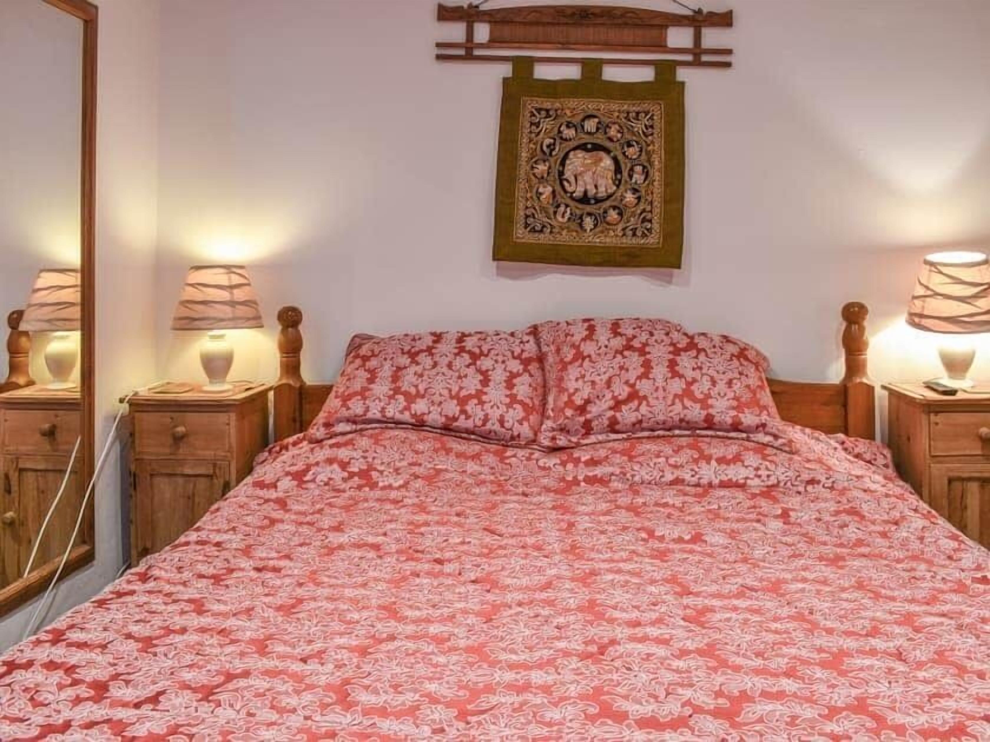 A bedroom with a bed, two bedside tables with lamps, a wooden headboard, and a wall hanging.