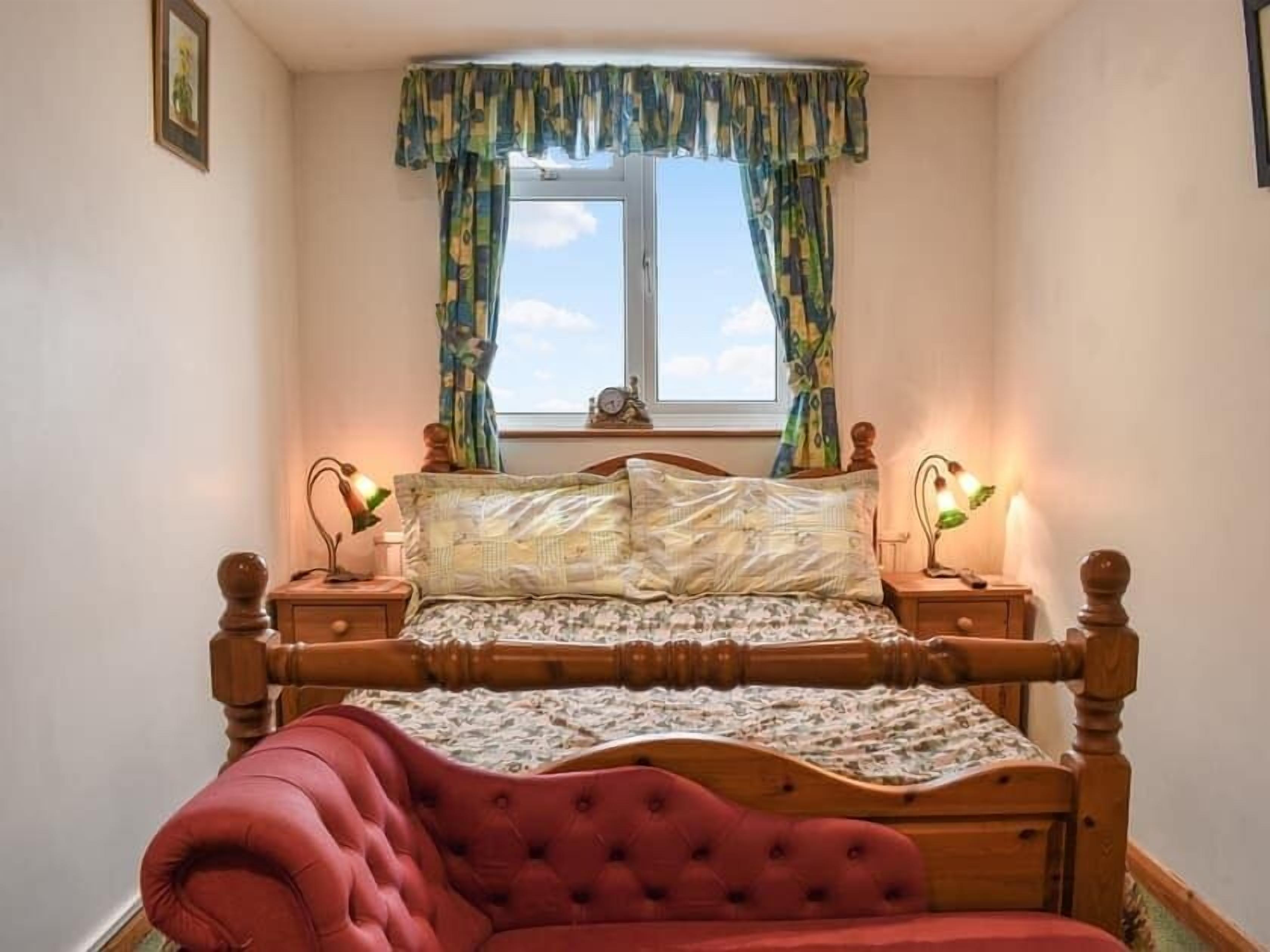 Une chambre avec un lit en bois, une literie à motifs floraux, un fauteuil recouvert de tissu rouge, deux tables de chevet avec des lampes et une fenêtre avec des rideaux.