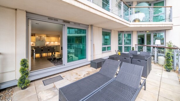 Une terrasse extérieure moderne comprenant un espace repas, des fauteuils de salon et une cuisine fermée par des parois vitrées.