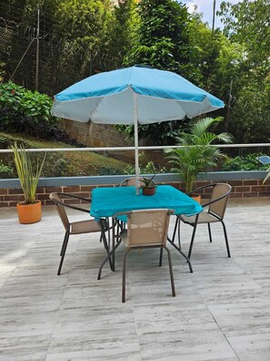 En udendørs patio med et bord og stole under en blå parasol, omgivet af plantekasser og en murstensmur.