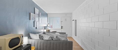 A compact living space with a grey sofa, a microwave, and a small wooden table. A white door and a window with blinds are visible.