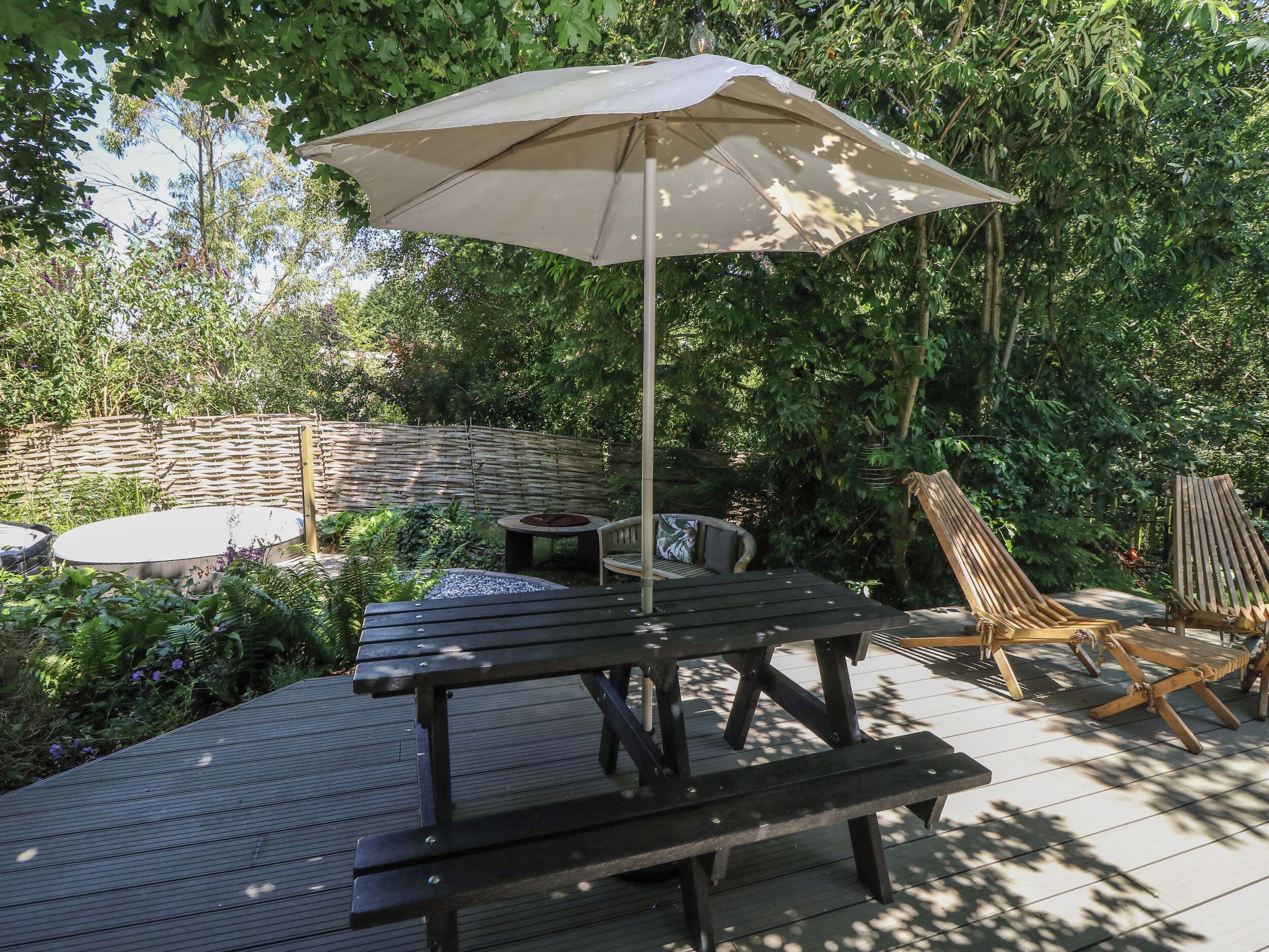 A wooden deck with a picnic table and an umbrella, surrounded by lush greenery and a woven fence.