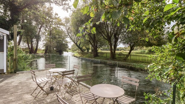 A riverside deck with folding chairs and a table, surrounded by lush greenery and trees. - The Happy Hut (Christchurch)