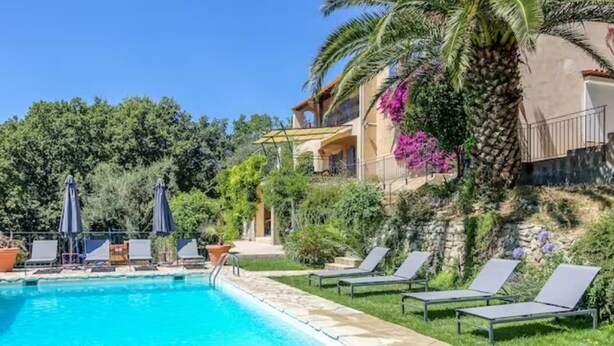 Piscina con sillones de descanso y sombrillas, rodeada de vegetación y un edificio con balcones.