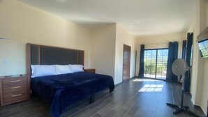 A bedroom with a bed, a nightstand, a dresser, a standing fan, and a large window with curtains. - Casa Camotlan (La Yesca)