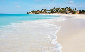A beach with clear turquoise water and white sand. There are buildings and palm trees in the background.