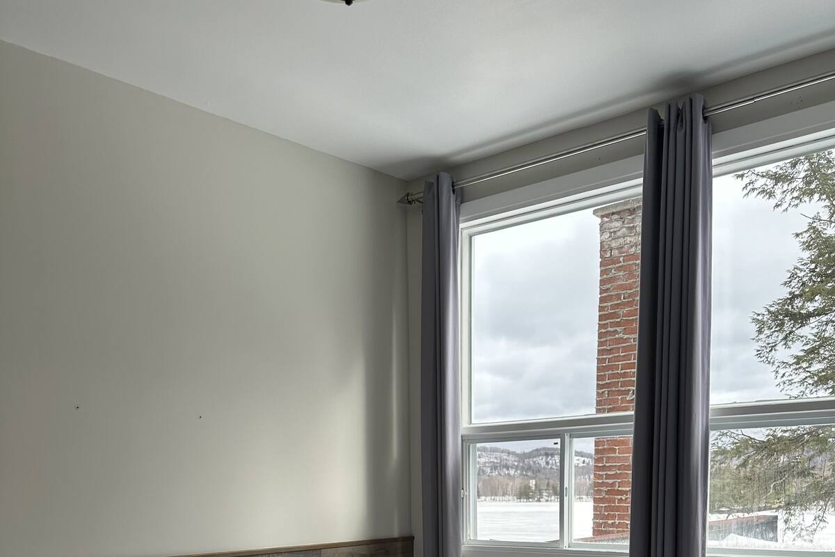 Ein Hotelzimmer mit Bett, einem Fenster mit Vorhängen und Blick auf ein Backsteingebäude und Bäume.